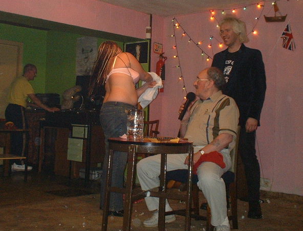 Gordon Cragg sits at the bingo table with Jagger standing behind dressed all in black looking like a body guard with a Jimi Saville wig on while a girl changes her top on stage